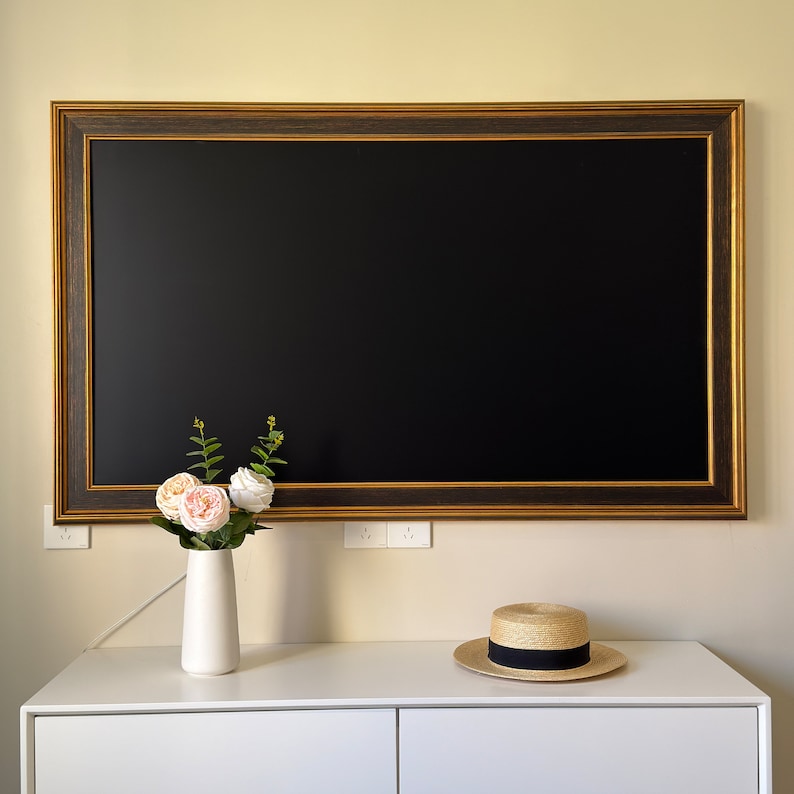 May include: A black framed mirror with a gold trim hangs on a white wall above a white dresser. A vase with pink flowers sits on the dresser. A straw hat with a black band sits on the dresser.