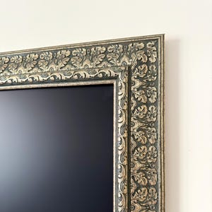 May include: A rectangular, ornate picture frame with a dark, matte center. The frame is dark green with gold accents and features a detailed, repeating leaf and scroll pattern. The frame is mounted on a white wall.