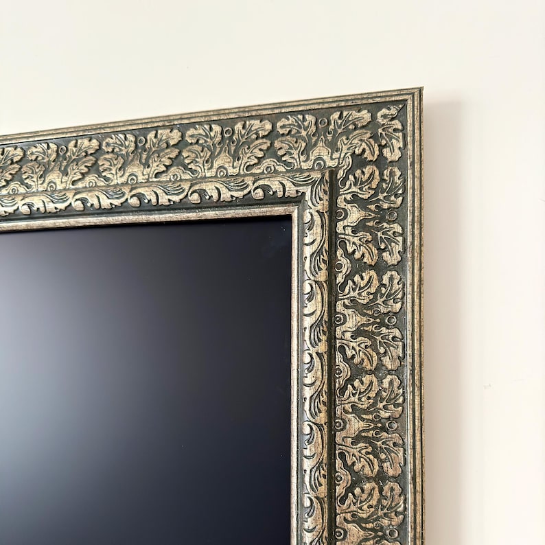 May include: A rectangular, ornate picture frame with a dark, matte center. The frame is dark green with gold accents and features a detailed, repeating leaf and scroll pattern. The frame is mounted on a white wall.