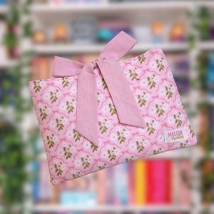 Könnte beinhalten: Eine rosa Stoff-Buchhülle mit floralem Muster und passender Schleife. Die Hülle hat ein sich wiederholendes Design aus weißen Blumen und grünen Blättern auf rosa Hintergrund. Das Etikett lautet "NALISA creates".
