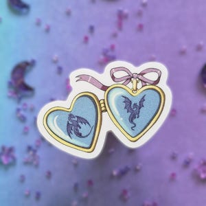 Könnte beinhalten: Ein Aufkleber, der ein offenes herzförmiges Medaillon mit Goldrand und hellblauem Innenraum zeigt. Auf beiden Seiten des Medaillons ist die Silhouette eines dunkelvioletten Drachen zu sehen. Eine hellviolette Schleife ist oben befestigt.