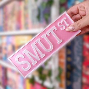 Könnte beinhalten: Ein rosa Straßenschild mit dem Text "SMUT ST" in weißen Blockbuchstaben. Das Schild hat einen erhabenen Rand und wird vor einem verschwommenen Hintergrund aus Büchern und Grün gehalten.