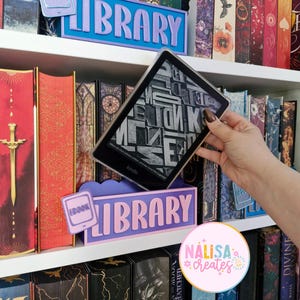 Può includere: Una libreria con insegne decorative che recitano "LIBRARY" e "EBOOK". Un e-reader Kindle mostra del testo. Gli scaffali sono pieni di libri colorati. Le insegne sono viola e blu.