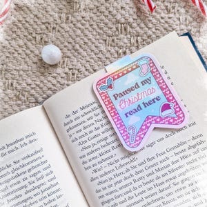 Könnte beinhalten: Ein farbenfroher Lesezeichen mit dem Text "Paused my Christmas read here" liegt auf einem offenen Buch. Das Lesezeichen hat ein rosa-blaues Design mit weihnachtlichen Illustrationen, darunter eine Zuckerstange und eine Weihnachtsmütze.