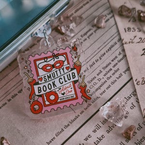 Könnte beinhalten: Ein klarer Acryl-Schlüsselanhänger mit einem rosa und roten Design. Der Schlüsselanhänger zeigt den Text "SMUTTY BOOK CLUB" mit einem roten Herzen und einem Paar roter Augenmasken. Rote Chilischoten und ein Donut sind ebenfalls enthalten.