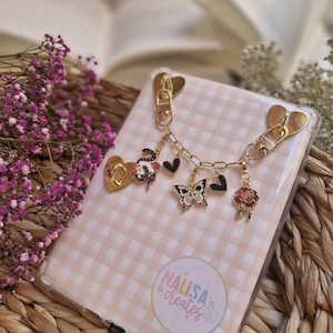 Könnte beinhalten: Eine transparente Handyhülle mit rosa Vichy-Muster, verziert mit einer goldenen Kette und Charms. Zu den Charms gehören eine Schlange, ein Schmetterling, Herzen und eine Rose. Die Hülle hat einen herzförmigen goldenen Verschluss. Der Text "NALISA *creates*" ist sichtbar.