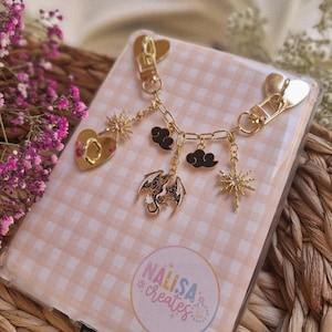 Könnte beinhalten: Transparente Handyhülle mit rosa-weißem Vichy-Muster. Eine goldfarbene Charm-Kette ist angebracht, mit einem Herz, Sonnenstrahlen, Wolken und einem Drachen. Der Text "NALISA creates" ist auf der Hülle aufgedruckt.