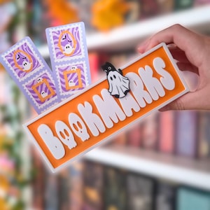 Könnte beinhalten: Ein rechteckiges orangefarbenes Schild mit weißer 3D-Beschriftung, die BOOKMARKS liest, mit einer kleinen schwarz-weißen Geisterkatzenfigur darauf. Dahinter sind zwei hellviolette rechteckige Lesezeichen mit orangefarbenen Rahmen und Geistermotiven zu sehen.