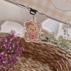 Könnte beinhalten: Ein klarer Acryl-Anhänger mit einem Cartoon-Teddybären, der ein rosa Buch liest. Der Bär hat eine rosa Schleife und ist an einem goldfarbenen Verschluss befestigt. Der Hintergrund umfasst einen geflochtenen Korb und lila Blumen.