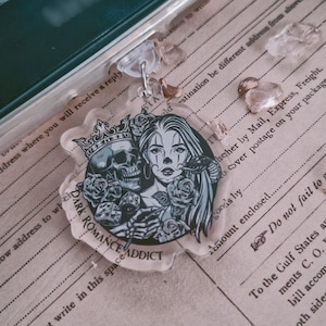 Op de afbeelding: Een heldere acryl sleutelhanger met een bloemen- en schedelontwerp. De sleutelhanger heeft het gezicht van een vrouw en een schedel met een kroon, omringd door rozen. De tekst "DARK ROMANCE ADDICT" is ook zichtbaar.