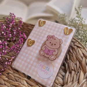 Könnte beinhalten: Eine transparente Handyhülle mit einem rosa-weiß karierten Muster. Die Hülle zeigt einen Cartoon-Bären, der ein Buch liest, und drei goldfarbene herzförmige Akzente. Die Hülle ist mit "NALISA CREATES" beschriftet.