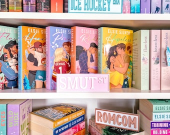 Könnte beinhalten: Ein weißes Bücherregal gefüllt mit verschiedenen Liebesromanen und vier rechteckigen Schildern. Die Schilder tragen den Text ICE HOCKEY ERA, SMUT ST, ROMCOM und NEW ADULT. Die Bücher sind in Reihen angeordnet, einige stehen aufrecht, andere liegen.