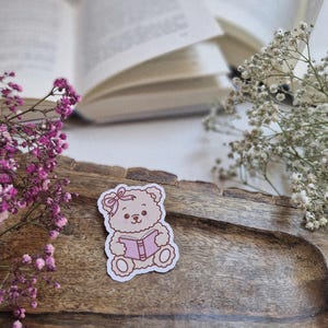 Könnte beinhalten: Ein Aufkleber mit einem niedlichen Cartoon-Teddybären, der ein rosa Buch liest. Der Bär hat eine rosa Schleife und ist braun umrandet. Der Aufkleber befindet sich auf einem Holztablett mit rosa und weißen Blumen und einem offenen Buch im Hintergrund.