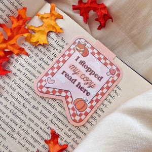 Könnte beinhalten: Ein Lesezeichen mit dem Text "I stopped my cozy read here" in einem rosa-weiß karierten Design, mit Illustrationen von Fuchs, Kürbis und Pilz. Das Lesezeichen befindet sich in einem offenen Buch, mit orangefarbenen und roten Herbstblättern verstreut.