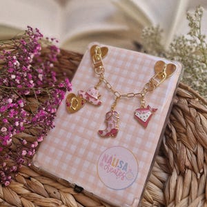 Könnte beinhalten: Eine transparente Handyhülle mit rosa Vichy-Muster und goldfarbenen Anhängern. Die Anhänger umfassen einen Cowboyhut, einen Stiefel, ein Herz und ein Halstuch. Auf der Hülle steht der Text "NALISA creates".