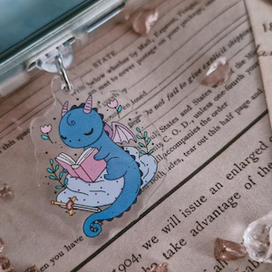 Op de afbeelding: Een heldere acryl sleutelhanger met een blauwe draak die een roze boek leest op een wolk. De draak heeft kleine hoorns en vleugels, met een zwaard en bloemen. De sleutelhanger is bevestigd aan een zilveren sluiting.