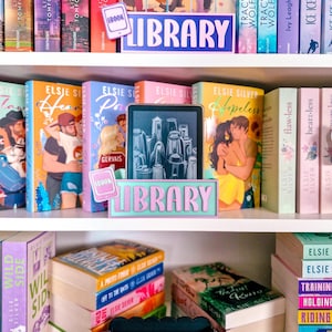 Könnte beinhalten: Ein weißes Bücherregal mit verschiedenen Büchern und drei rechteckigen Haltern. Zwei Halter sind hellblau mit rosa Schriftzug LIBRARY und einem E-Book-Symbol. Ein Halter ist schwarz mit kupferfarbenem Schriftzug LIBRARY und E-Book-Symbol.