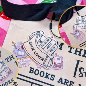 Könnte beinhalten: Eine beigefarbene Tragetasche mit schwarzem Griff und einem rosa-weißen Blumenmuster. Die Tasche hat Aufkleber mit Buchmotiven und den Texten "Book Lover", "Talk Bookish to Me" und "Bookish Vibes Only".