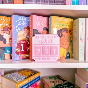 Könnte beinhalten: Ein Bücherregal mit Büchern mit farbenfrohen Einbänden. Ein rosa Bücherzähler mit dem Text "From the Library of Izabell" und der Zahl "231" steht im Mittelpunkt. Die Bücher sind von Elsie Silver und anderen Autoren.