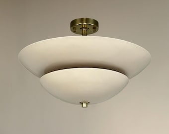 Pode incluir: Uma luminária de teto semi-embutida com dois abajures sobrepostos em forma de taça, na cor branco-sujo e acabamento fosco. A luminária é conectada por uma haste central a uma canopla circular em tom de latão e um pequeno remate de latão.