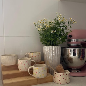 May include: Three ceramic mugs and two small bowls with hand-painted colourful flower patterns, a tall white ribbed vase filled with white and yellow daisies, and a pink stand mixer with a stainless steel bowl on a white kitchen counter.