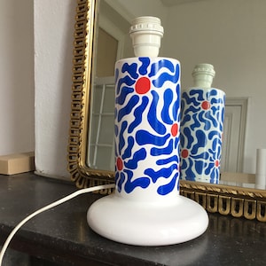 Op de afbeelding: Een witte keramische tafellamp met een abstract bloemenmotief in blauw en rood. De lamp heeft een wit snoer en een witte voet.