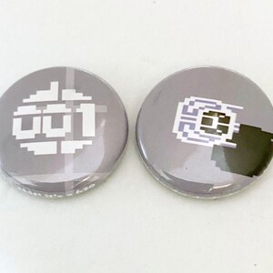 May include: Two round grey button badges with white pixelated designs. The first badge features the word "001" in a pixelated font. The second badge features a pixelated design of a circuit board.