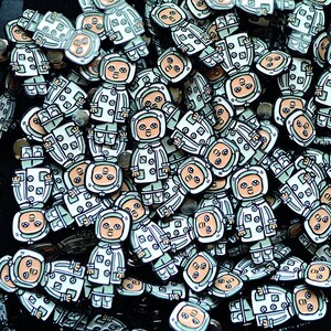 May include: A large pile of enamel pins featuring a cartoon character wearing a white spacesuit with a green jacket. The character has a round head with a light brown face and black eyes. The pins are all different colours and have a variety of designs.