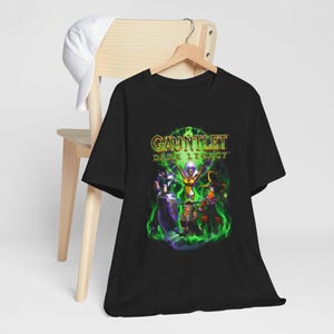 May include: A black short-sleeve t-shirt featuring a graphic print of the Gauntlet Dark Legacy video game logo and characters surrounded by green flames. The shirt is displayed on a wooden hanger, with a white baseball cap hanging nearby.