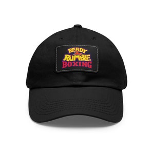 May include: Black baseball cap with a curved brim. The cap features a rectangular patch with the words "READY 2 RUMBLE BOXING" in yellow, red, and blue. The text is outlined in white. The cap is made of a soft material.