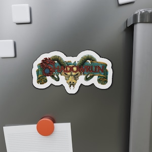 Könnte beinhalten: Ein Magnet mit einem stilisierten Schädel, geschwungenen Hörnern und dem Wort "SHADOWRUN" auf einem Banner. Das Design verwendet Rot-, Blau- und Goldtöne mit einem weißen Rand. Der Magnet ist auf einer grauen Oberfläche befestigt.