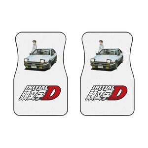 May include: Pair of white car floor mats featuring a light blue car with a person standing next to it. The mats have the text "INITIAL D" in red and black. The car has yellow headlights.