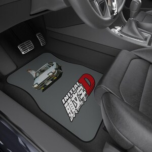 May include: Gray car floor mats with a black border featuring a graphic of a car and the text "INITIAL D" in white and red. The mats are shown inside a car.