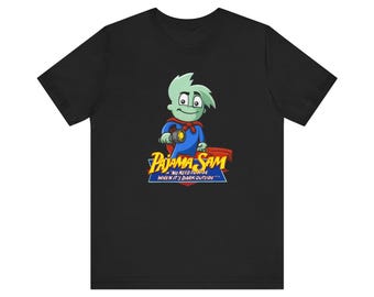 May include: Black t-shirt featuring a cartoon character named Pajama Sam. The character is green and wearing a blue and red superhero costume. The text on the shirt reads "Pajama Sam - No need to hide when it's dark outside."