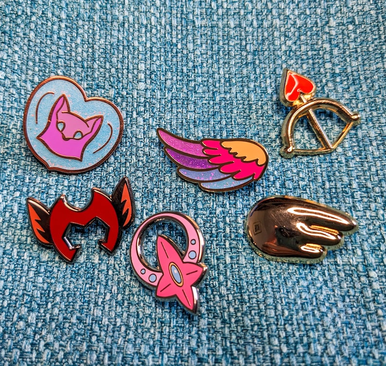 May include: A set of ten enamel pins featuring various designs, including a heart with a cat face, a wing, a bow and arrow, a star, and a hand. The pins are all gold-colored with pink, red, and purple accents.