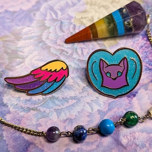 May include: Three enamel pins with a rainbow wing, a purple cat in a blue heart, and a rainbow crystal pendulum. The pins are on a purple and blue background.