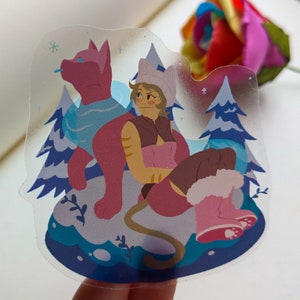 Puede incluir: Una pegatina transparente con un gato rosa que lleva una bufanda azul y una mujer con cabello castaño que lleva un sombrero rosa y botas. La mujer está sentada en una colina cubierta de nieve con un fondo azul. La pegatina tiene un tema invernal.