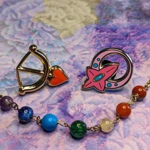 May include: Two enamel pins, one with a gold bow and arrow with a red heart, and the other with a pink flower in a silver crescent moon. The pins are on a purple and pink background with a beaded necklace.