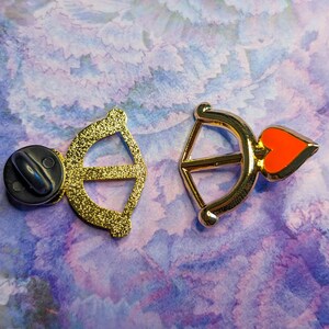 May include: Two gold-tone metal pins, one shaped like an arrow with a red heart and the other shaped like a bow.