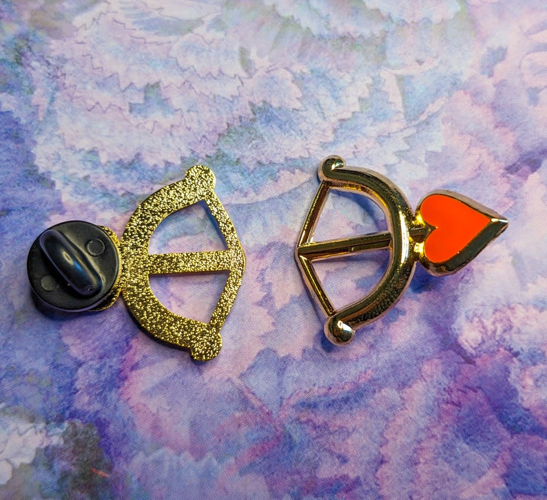 May include: Two gold-tone metal pins, one shaped like an arrow with a red heart and the other shaped like a bow.