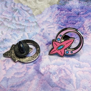 May include: Two enamel pins, one with a pink star and a silver crescent moon, the other with a silver crescent moon and a black pin back.