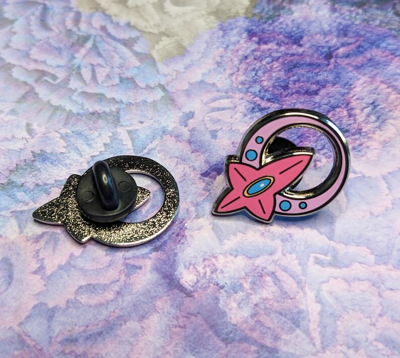 May include: Two enamel pins, one with a pink star and a silver crescent moon, the other with a silver crescent moon and a black pin back.