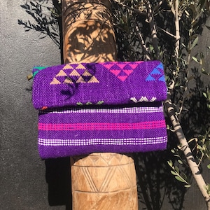 Peut inclure: Une pochette tissée violette avec un motif géométrique rose, bleu et beige. La pochette a un rabat et est faite d'un tissu texturé. Le sac repose sur un poteau en bois sculpté, avec de la verdure en arrière-plan.