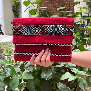 Peut inclure: Une pochette rectangulaire rouge avec un motif tissé. La pochette présente un motif géométrique noir, blanc et bleu sur le dessus. La pochette est tenue devant un fond de plantes vertes et un tronc d'arbre.