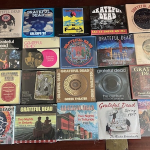 May include: A collection of Grateful Dead album covers and concert posters. The covers feature various designs, including skulls, band logos, and concert locations. The collection includes albums from the 1980s and earlier, with vibrant colors and unique artwork.