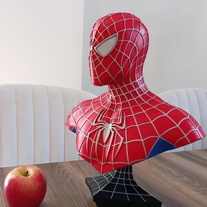 May include: A red and blue Spider-Man bust figurine with a black webbed base. The bust is on a wooden table with a red apple in the foreground.