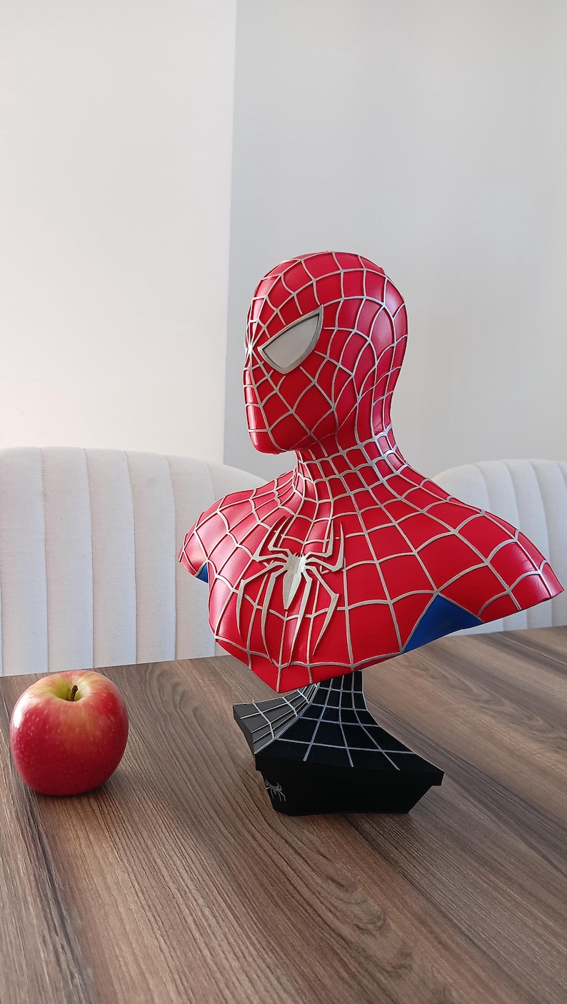 May include: A red and blue Spider-Man bust figurine with a black webbed base. The bust is on a wooden table with a red apple in the foreground.