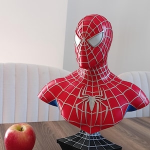 May include: A red and blue Spider-Man bust with a black webbed base. The bust is on a wooden table with a red apple.