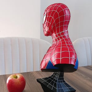 May include: A red and blue Spider-Man bust figurine with a black web-shaped base. The bust is on a wooden table with a red apple.