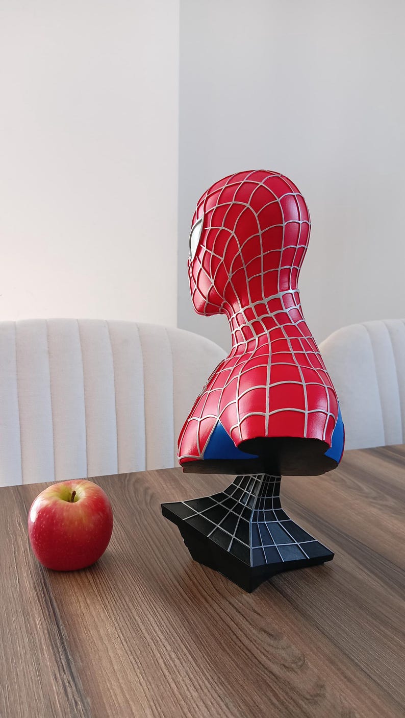 May include: A red and blue Spider-Man bust figurine with a black web-shaped base. The bust is on a wooden table with a red apple.